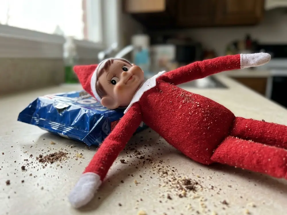 Toy elf doll with a round belly lying among cookie crumbs and an empty package