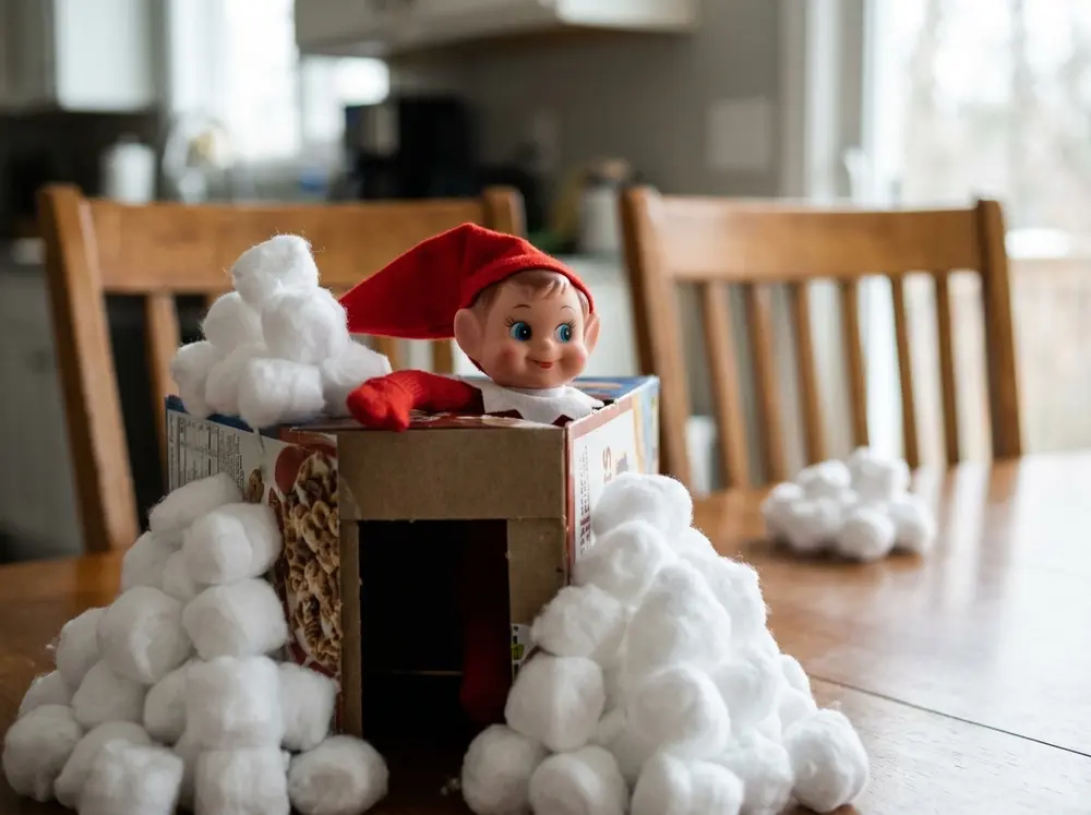 Toy elf doll behind a cereal-box fort surrounded by cotton-ball snowballs