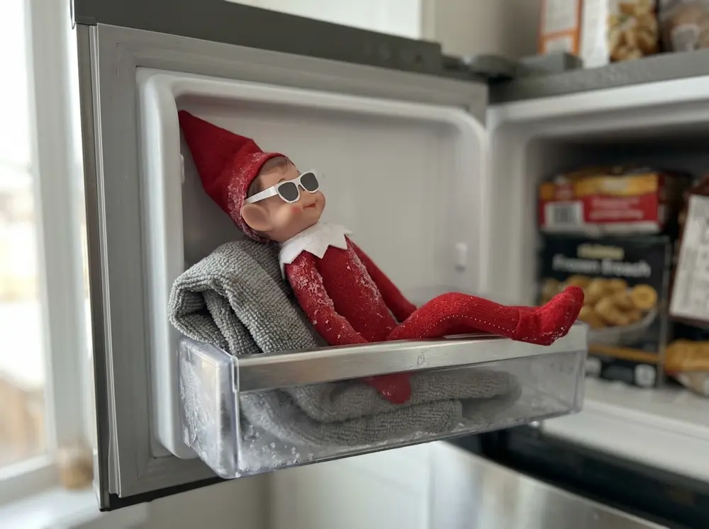 Toy elf doll lounging on a facecloth towel in the freezer wearing paper sunglasses