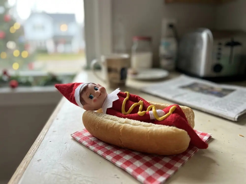 Toy elf doll sleeps tucked into a hot-dog bun like a sleeping bag, squiggle of (drawn or real) mustard as a blanket