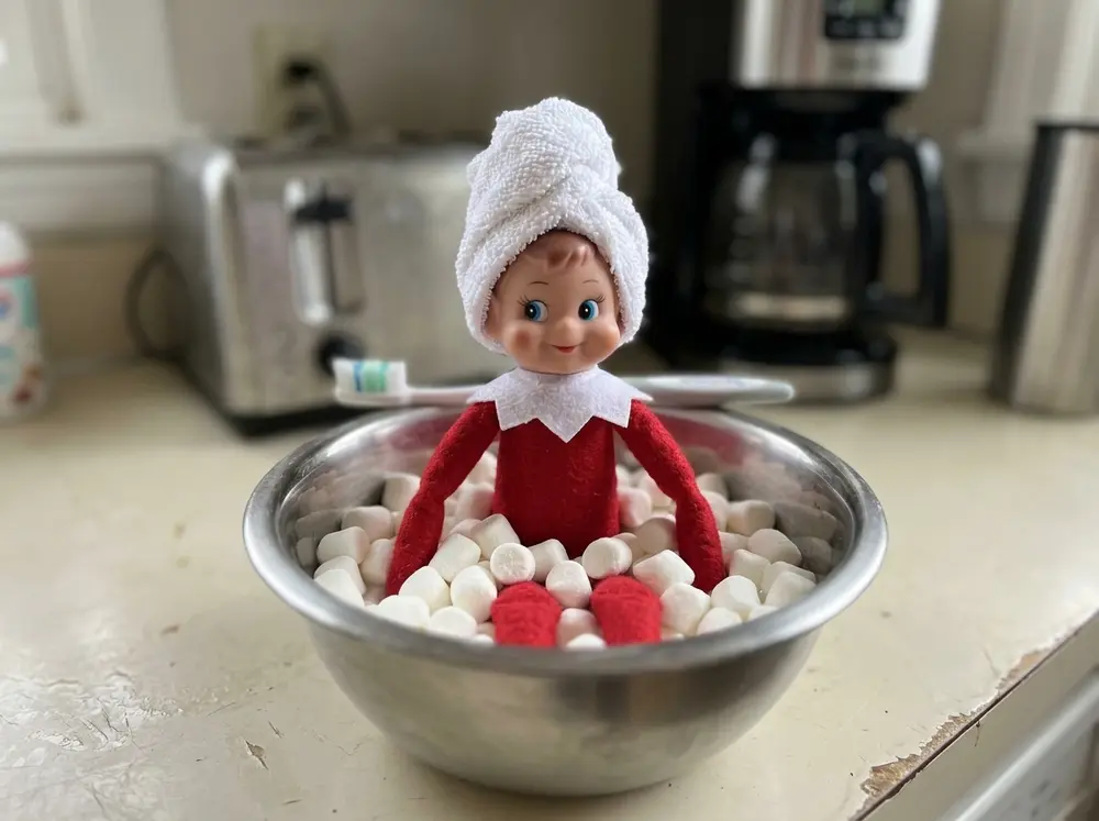 Toy elf doll bathing in a mixing bowl of mini marshmallows with a washcloth turban