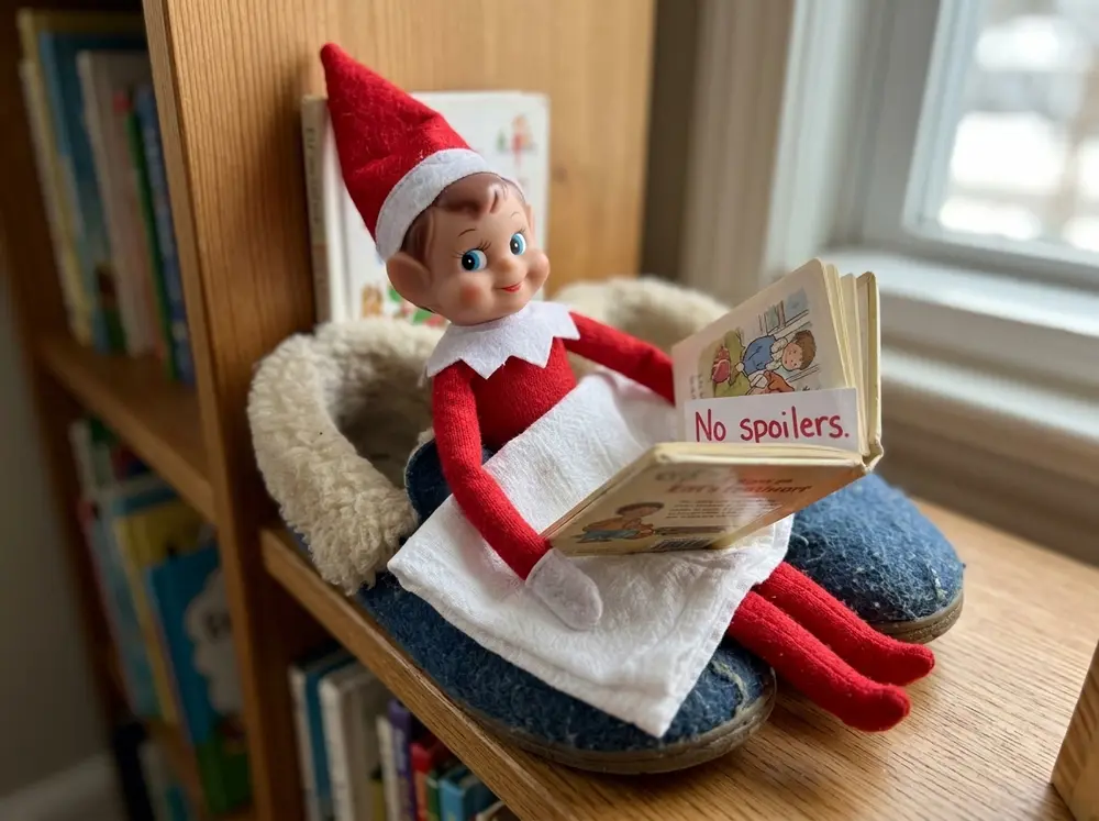 Toy elf doll builds a nook from a slipper and a facecloth blanket, mid-way through the child's favourite book with a bookmark note: 'No spoilers.'
