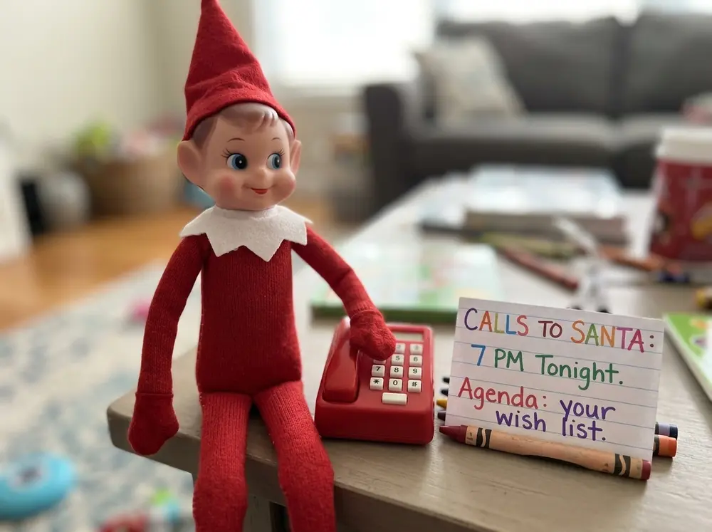 Toy elf doll mans a toy phone with a sign-up sheet: 'Calls to Santa, 7 PM tonight