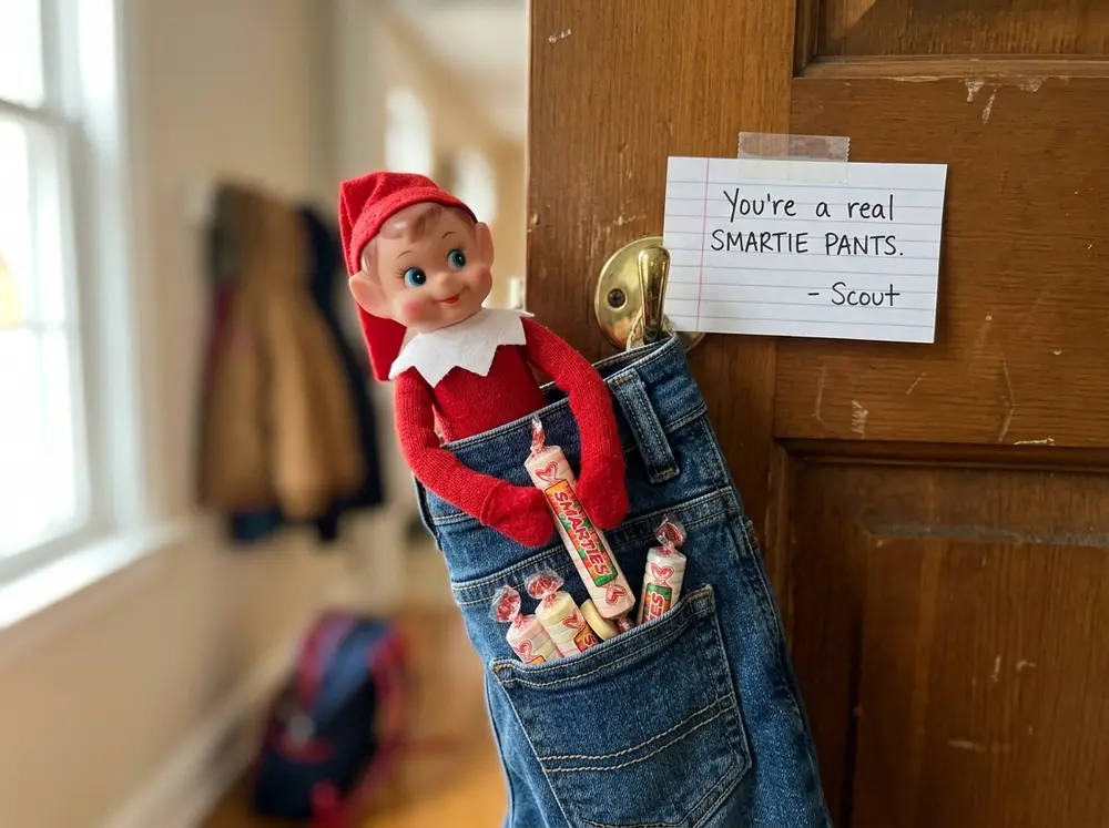 Toy elf doll tucks candy into a pair of the kid's pants hanging on the door with a note: 'You're a real SMARTIE PANTS.'
