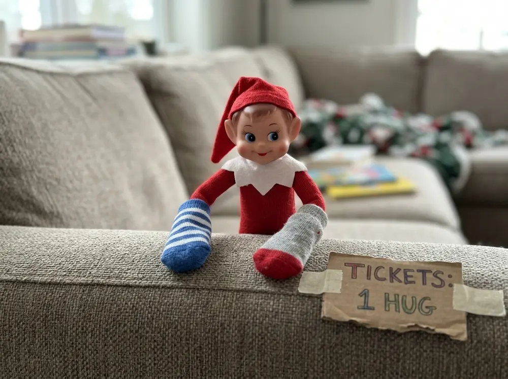 Toy elf doll runs a puppet show from behind the couch arm with two socks on its hands and a 'TICKETS: 1 HUG' sign