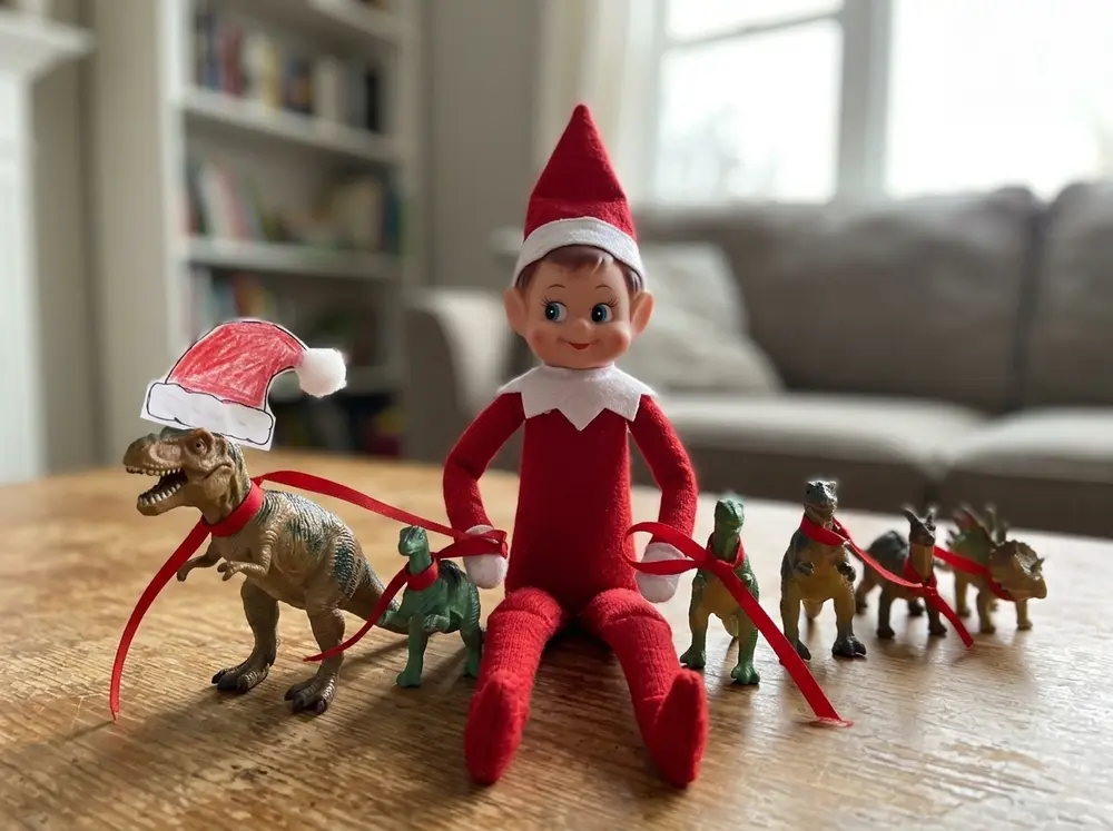 Toy elf doll 'tames' the toy dinosaurs: lined up, ribbon lassos on, one dino wearing a Santa hat drawn on paper